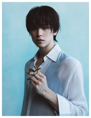 A young man with dark tousled hair wearing a white shirt holding a butterfly on his finger against a light blue background.