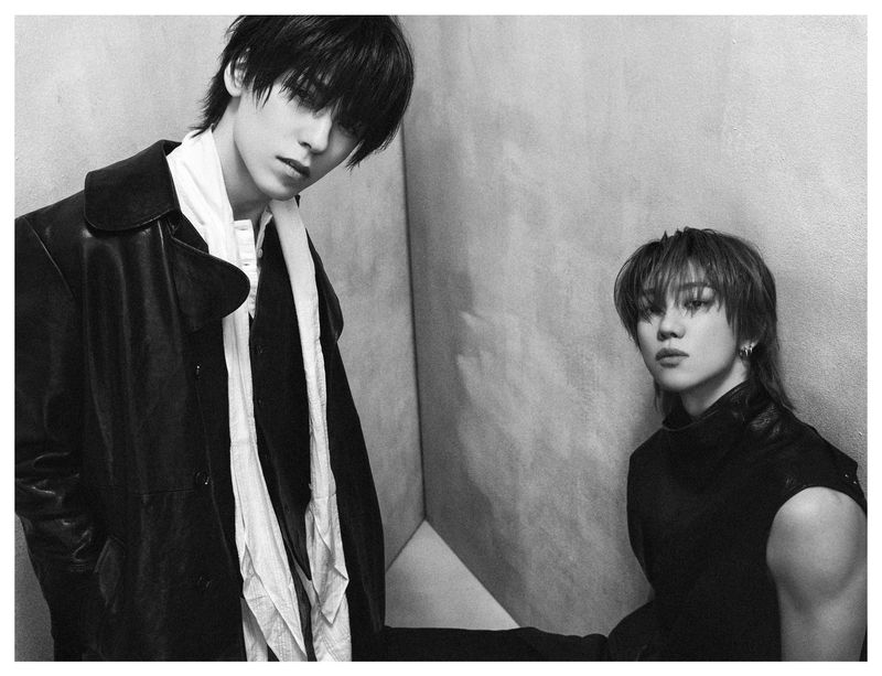 Black and white portrait of two men in dark leather jackets posing against a textured corner wall