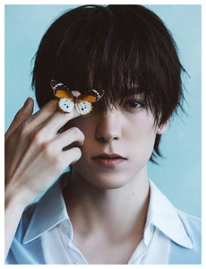 Portrait of young man with dark hair holding a butterfly on his finger near his eye against a light blue backdrop.