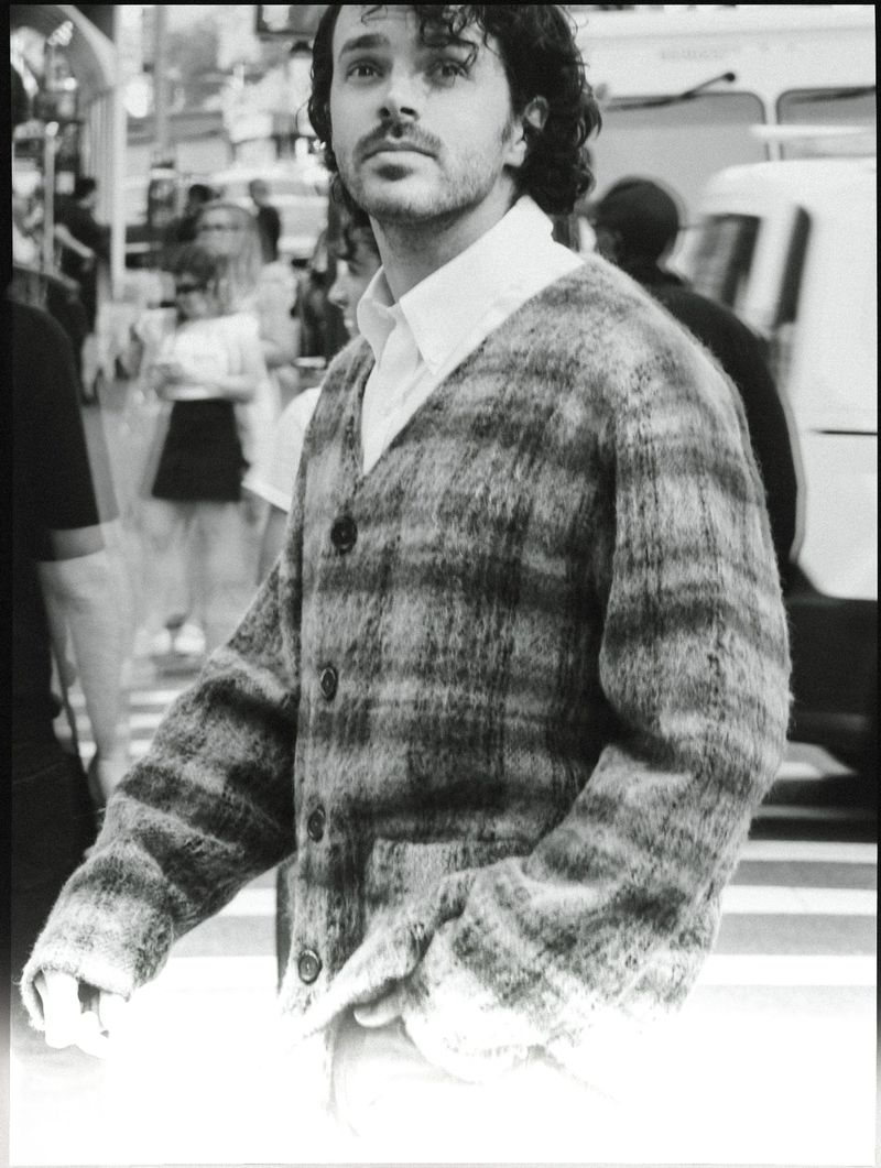 Black and white portrait of an adult male with curly hair, wearing a patterned cardigan sweater on a city street with blurred figures.