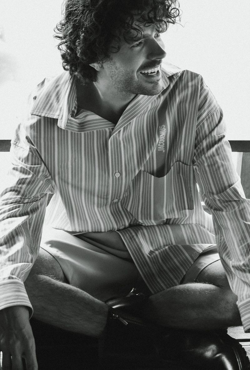 Monochrome portrait of a smiling man with curly hair wearing a striped shirt and shorts sitting cross-legged indoors.