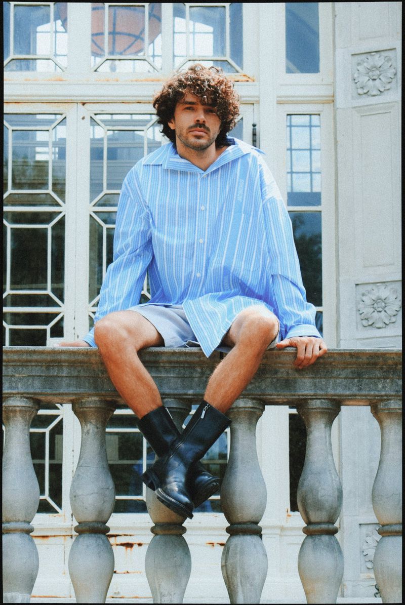Adult man with curly hair poses outdoors on a stone ledge wearing boots, shorts, and a striped shirt in an eye-level portrait shot.