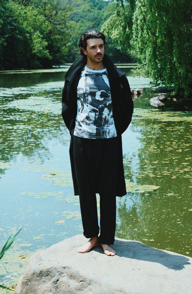 Harry Richardson standing barefoot on a rock beside a pond wearing a black coat, graphic t-shirt, and black trousers.