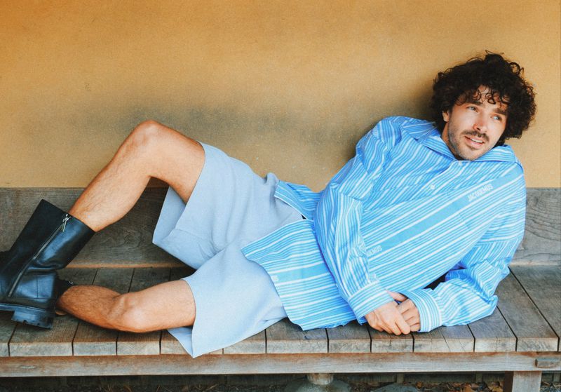 Man with curly hair in a striped shirt and shorts reclines on a wooden surface in an indoor environment, posing and looking at the camera.