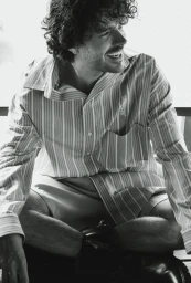 Harry Richardson sitting cross-legged wearing an oversized striped shirt in a black and white editorial portrait.