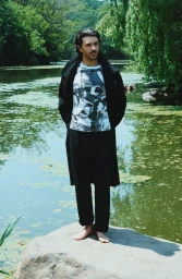 Harry Richardson standing barefoot on a rock beside a pond wearing a black coat, graphic t-shirt, and black trousers.