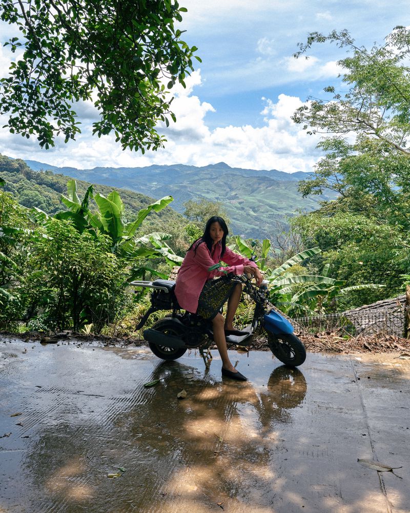 Model wearing a pink shirt seated on a small blue motorbike on an outdoor mountain path with lush green vegetation
