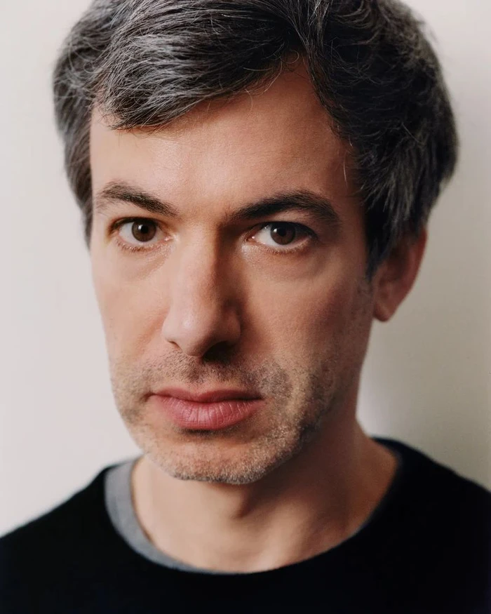 Portrait of Nathan Fielder with short dark hair and stubble wearing a black sweater against a plain white background