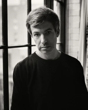 Portrait of Nathan Fielder wearing a dark crewneck sweater standing in front of a multi-pane window.