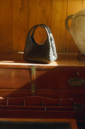 Black woven leather handbag resting on a polished wooden cabinet against wooden wall panels.