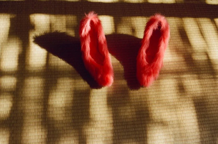 Pair of bright red fluffy faux-fur shoes resting on a textured woven mat under dramatic striped shadows.