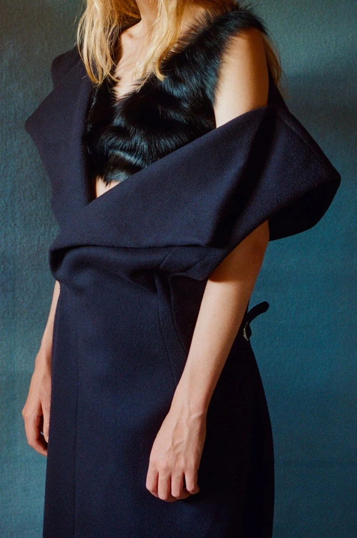 Model wearing a dark structured wool coat and black faux fur top against a textured dark teal background.
