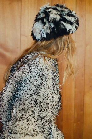 Model with long blonde hair wearing a textured black and white fluffy coat and matching hat against wood paneling