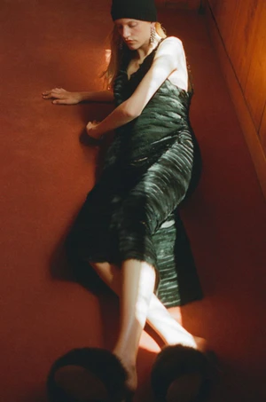 Model wearing a black dress and beanie, reclining on a red carpeted floor with warm sunlight streaming across the scene.