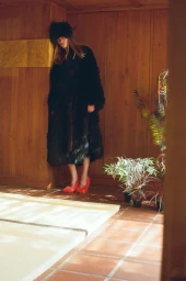 Model in black fur coat and hat with red shoes leaning against wood panelled wall