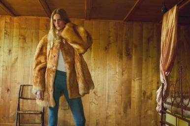 Model wearing a fuzzy brown coat and blue trousers inside a wooden paneled room with a rustic bed.