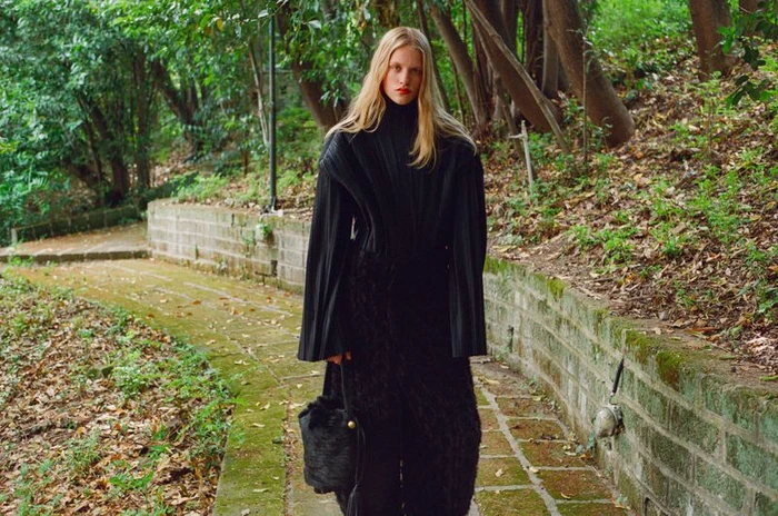 Model in a black pleated turtleneck and textured black skirt holding a handbag on a park path