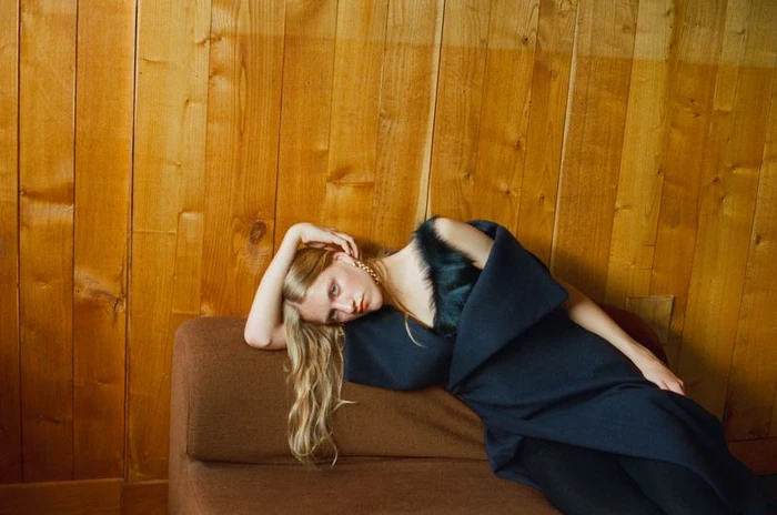 Model with long blonde hair reclining on a brown sofa against wood paneling wearing a dark blue dress with fur trim