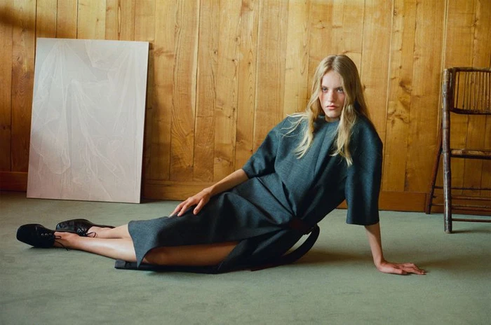 Model seated on a green carpet in a dark grey dress against a wooden panel wall