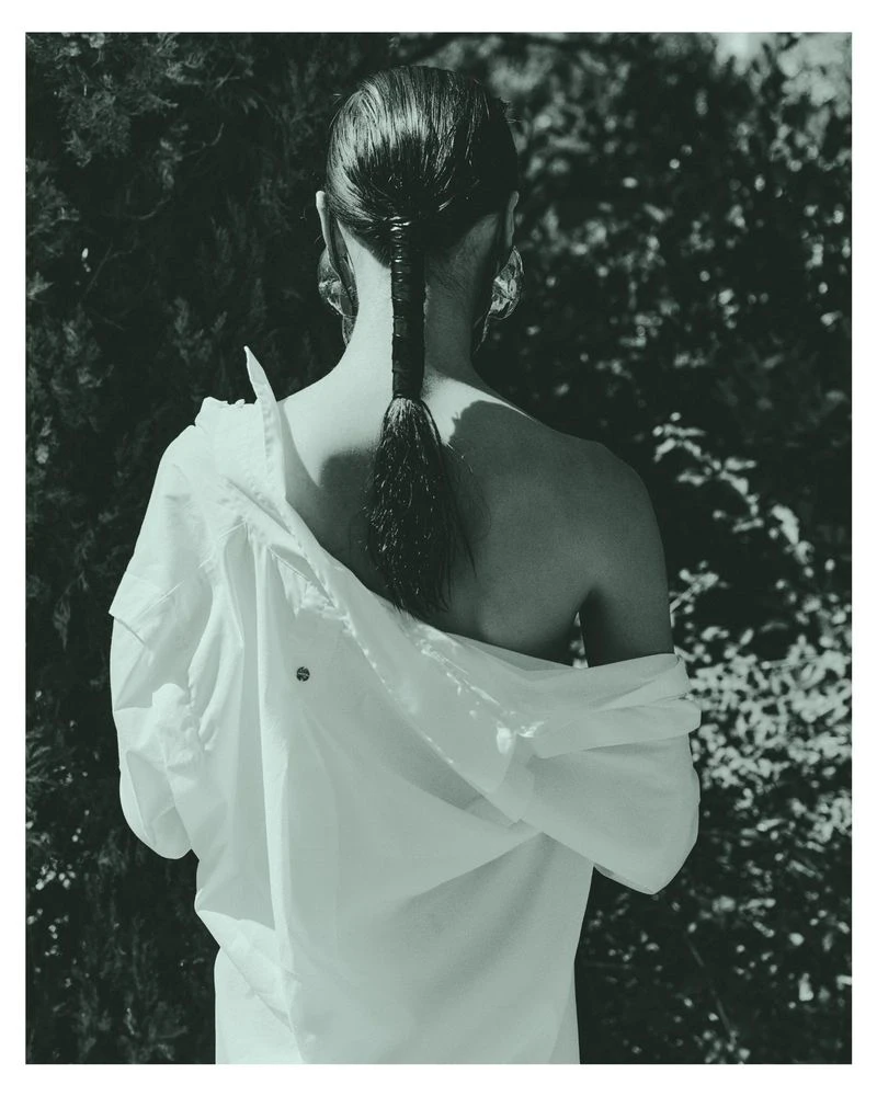 Model Dominika Pikula seen from behind wearing an off-the-shoulder white shirt against a dark leafy background for Grazia Germany.