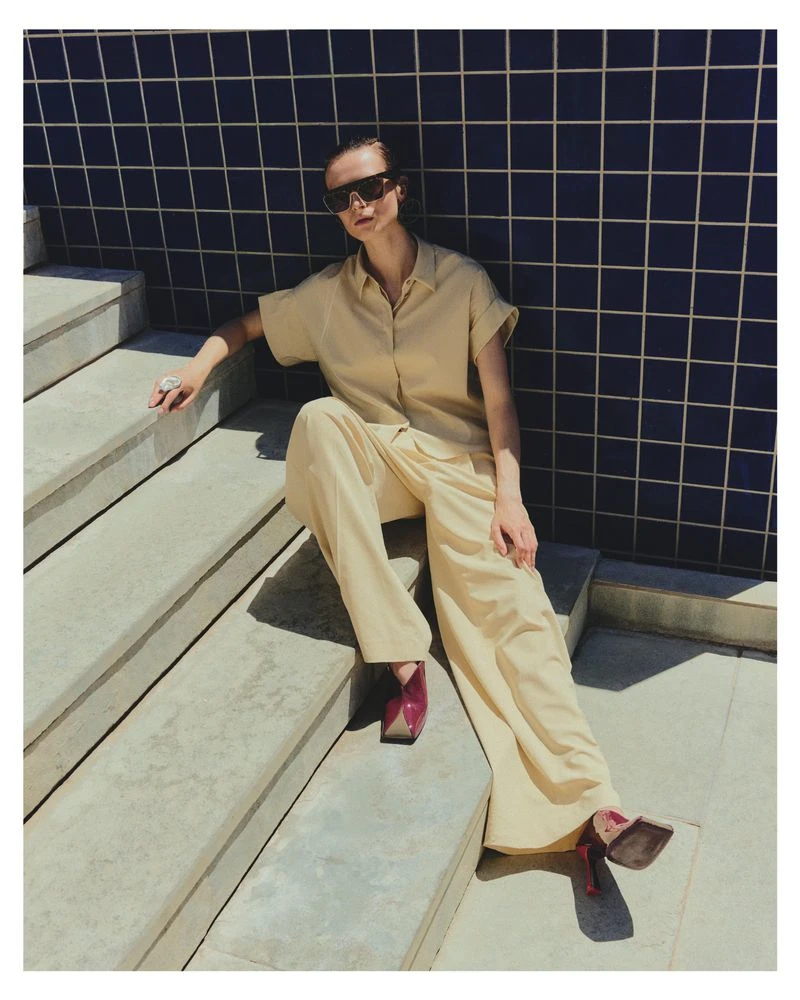 Model Dominika Pikula seated on concrete outdoor stairs wearing cream button-up shirt and wide trousers with dark sunglasses