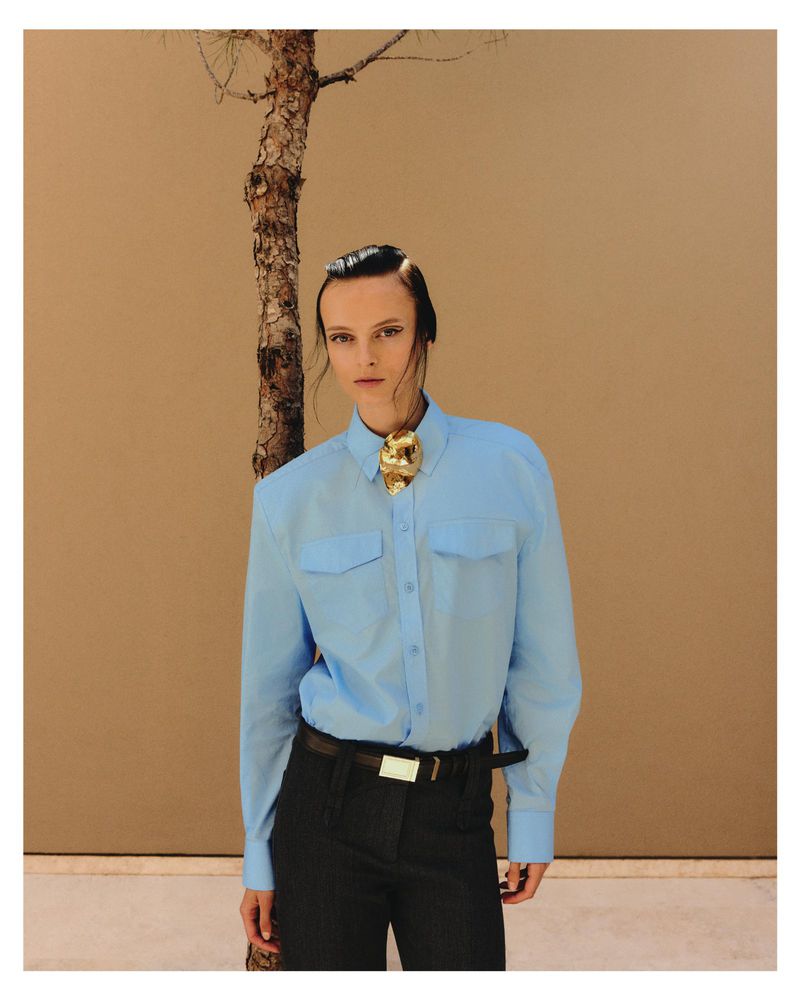 Model Dominika Pikula wearing a light blue button-down shirt and dark trousers against a plain wall with a thin tree trunk