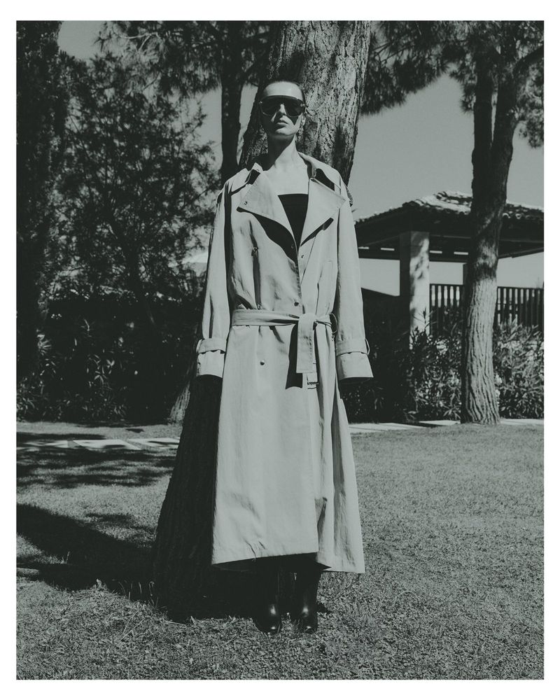 Model Dominika Pikula wearing sunglasses and a long belted trench coat standing outdoors on grass under trees for Grazia Germany.