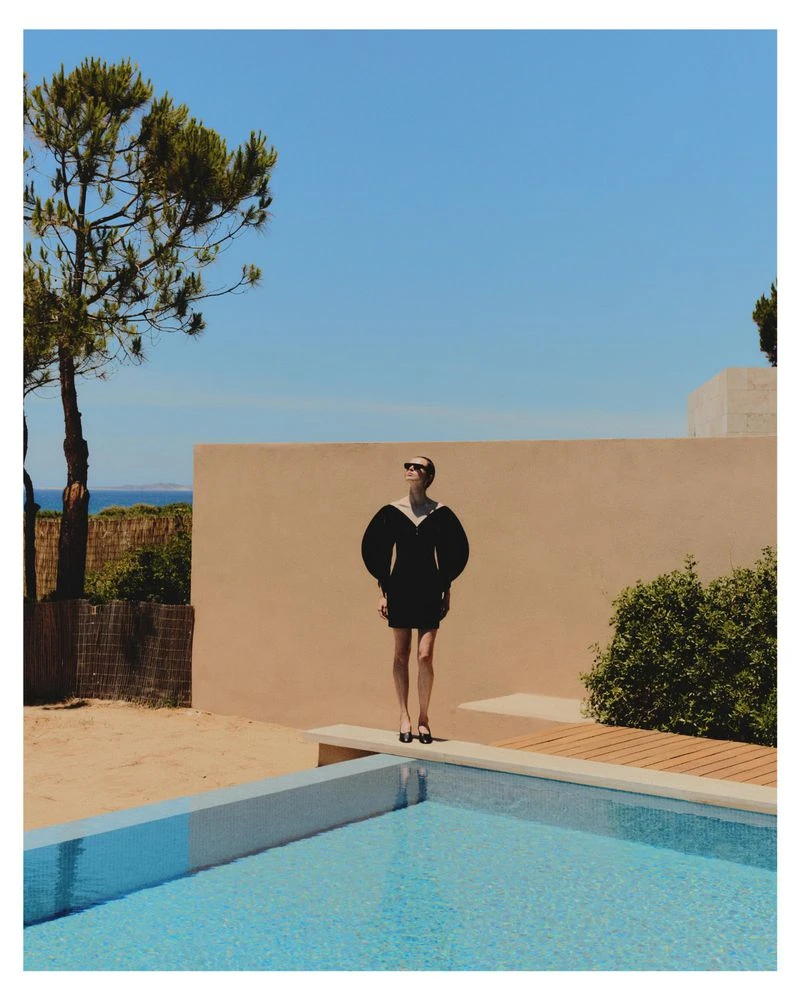 Model wearing a black dress by a swimming pool under clear blue sky for Grazia Germany