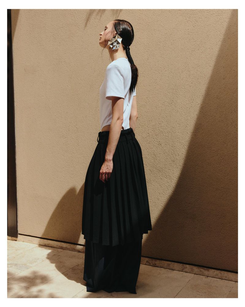 Model Dominika Pikula wearing a white top and black pleated skirt against a plain wall for Grazia Germany.