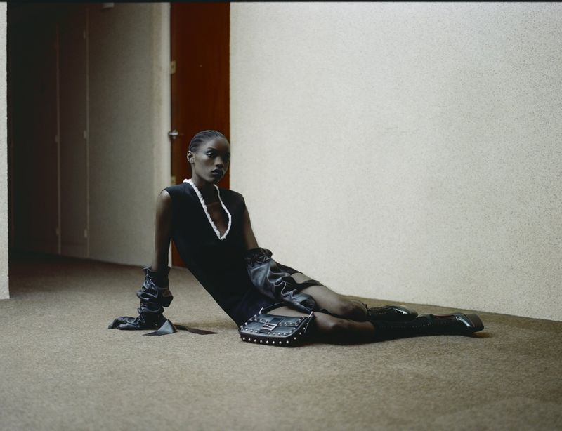 Black model wearing sleeveless black dress and long leather gloves seated on carpeted floor in a hallway.