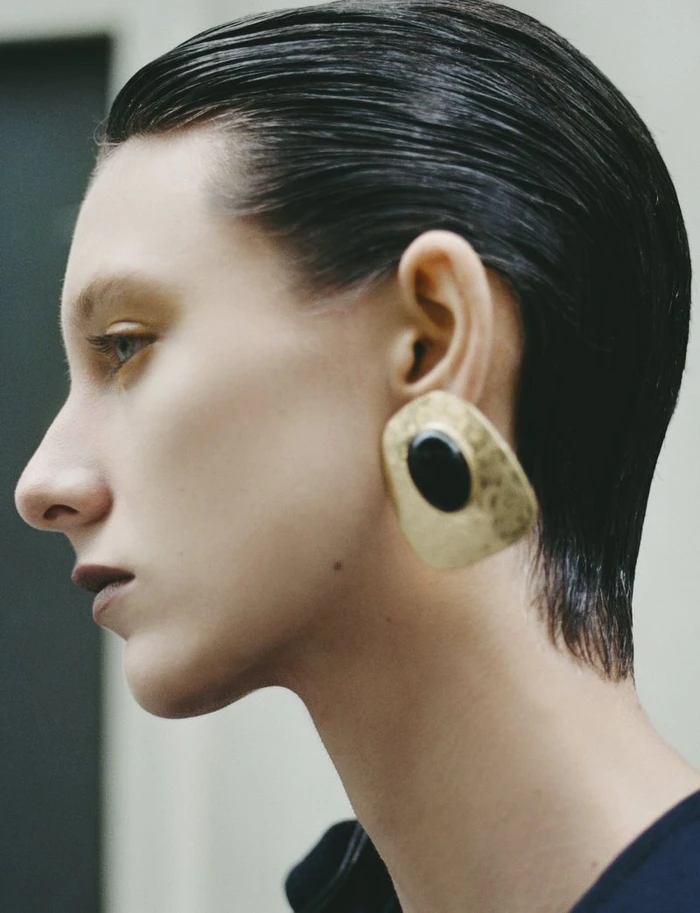Profile portrait of a woman with slicked-back dark hair wearing a large gold statement earring with a black center