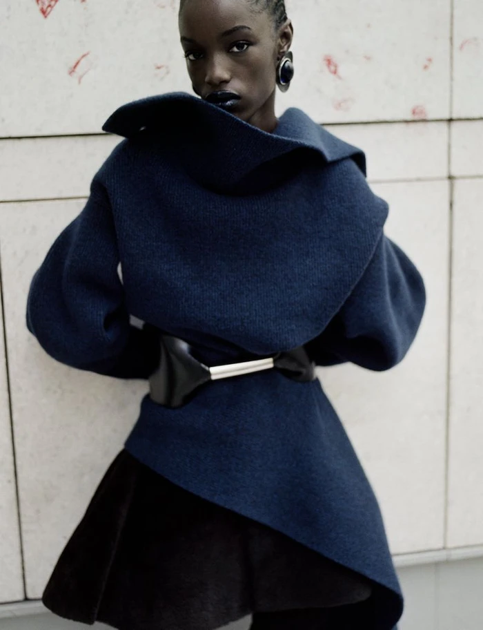Model in oversized dark blue sweater and black skirt posing against a stone wall with red markings