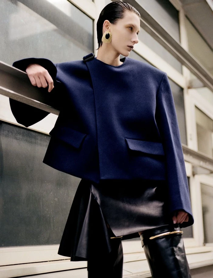 Model with slicked-back hair wearing a structured blue jacket and leather skirt leaning against an exterior building railing