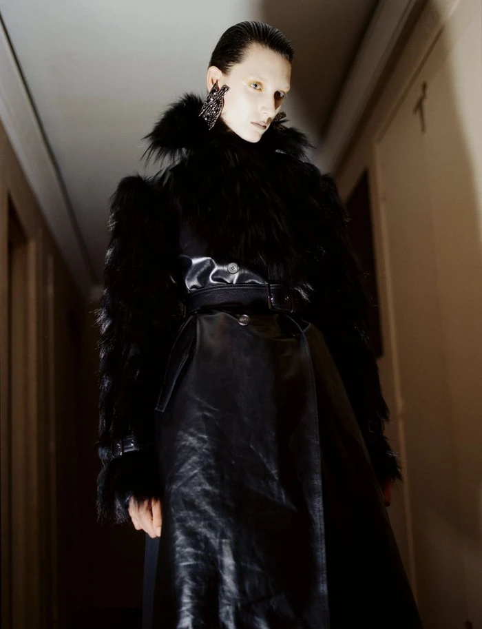 Model wearing a black faux-fur trimmed coat and large statement earrings in an indoor hallway setting.
