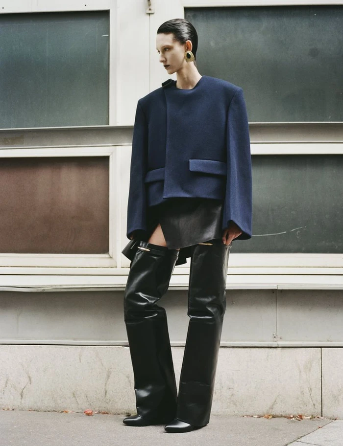 Model in blue structured jacket and black leather thigh-high boots against a building exterior.