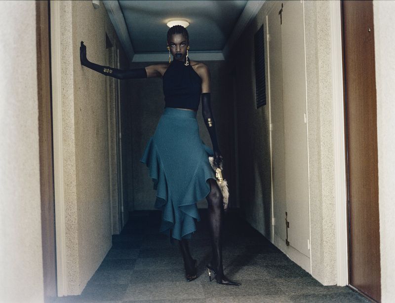 Model standing in a narrow hallway wearing a black top, teal ruffled skirt, and long black gloves