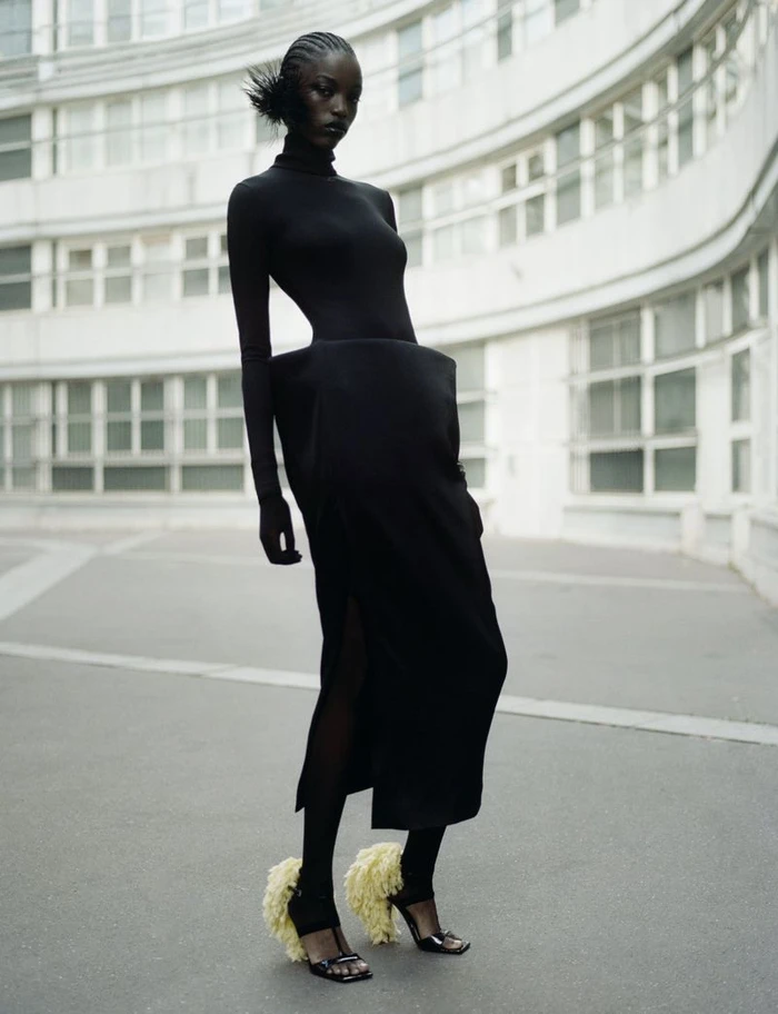Model in black sculptural dress and feathered heels posing outdoors before curved white modernist building architecture