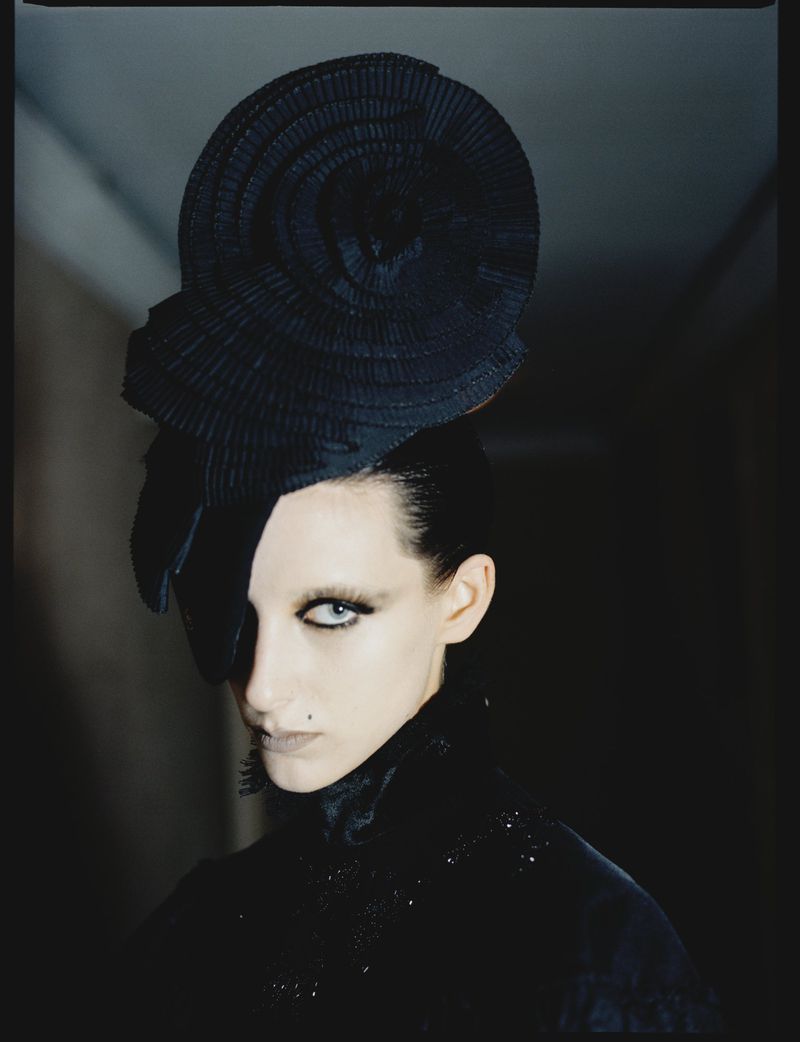 Model wearing a large sculptural black hat and dark clothing against a dim background for the Where in Paris project.