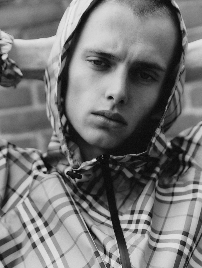 Monochrome portrait of a young adult male with a neutral expression, wearing a plaid hooded jacket and posing against a brick wall.