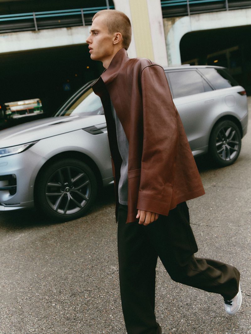 Male model in brown leather jacket walks past a silver Range Rover in an urban setting, showcasing automotive fashion and modern design.