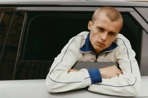 Close-up of a young man with a shaved head leaning his arms on the window frame of a car, wearing a white jacket with blue piping.