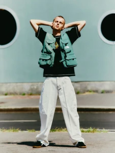 Model Elias Monstrey standing outdoors wearing a green tactical utility vest, black t-shirt, and wide-leg white trousers.