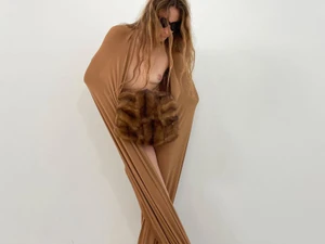 Model in sunglasses wearing a wrapped brown fabric garment and holding a textured fur muff against a white background.