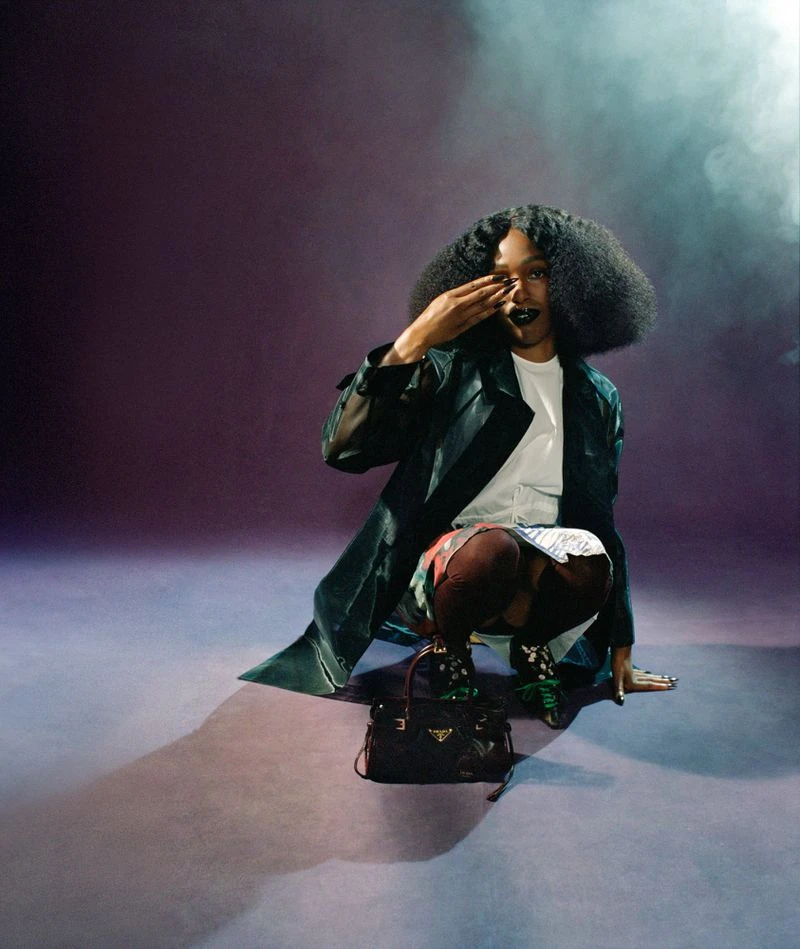 John Glacier crouched in a dark green coat and white t-shirt next to a black handbag against a purple studio backdrop