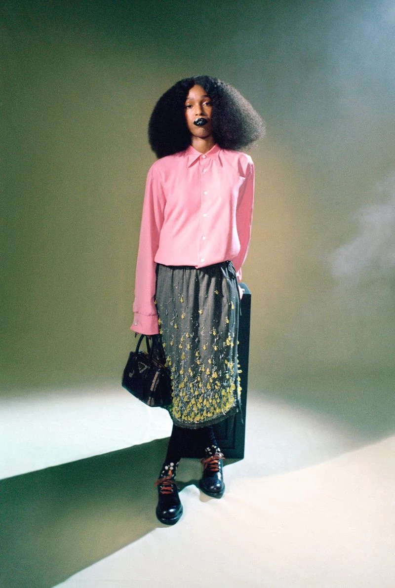 John Glacier in a pink shirt and grey skirt holding a black handbag against a studio backdrop for i-D Magazine