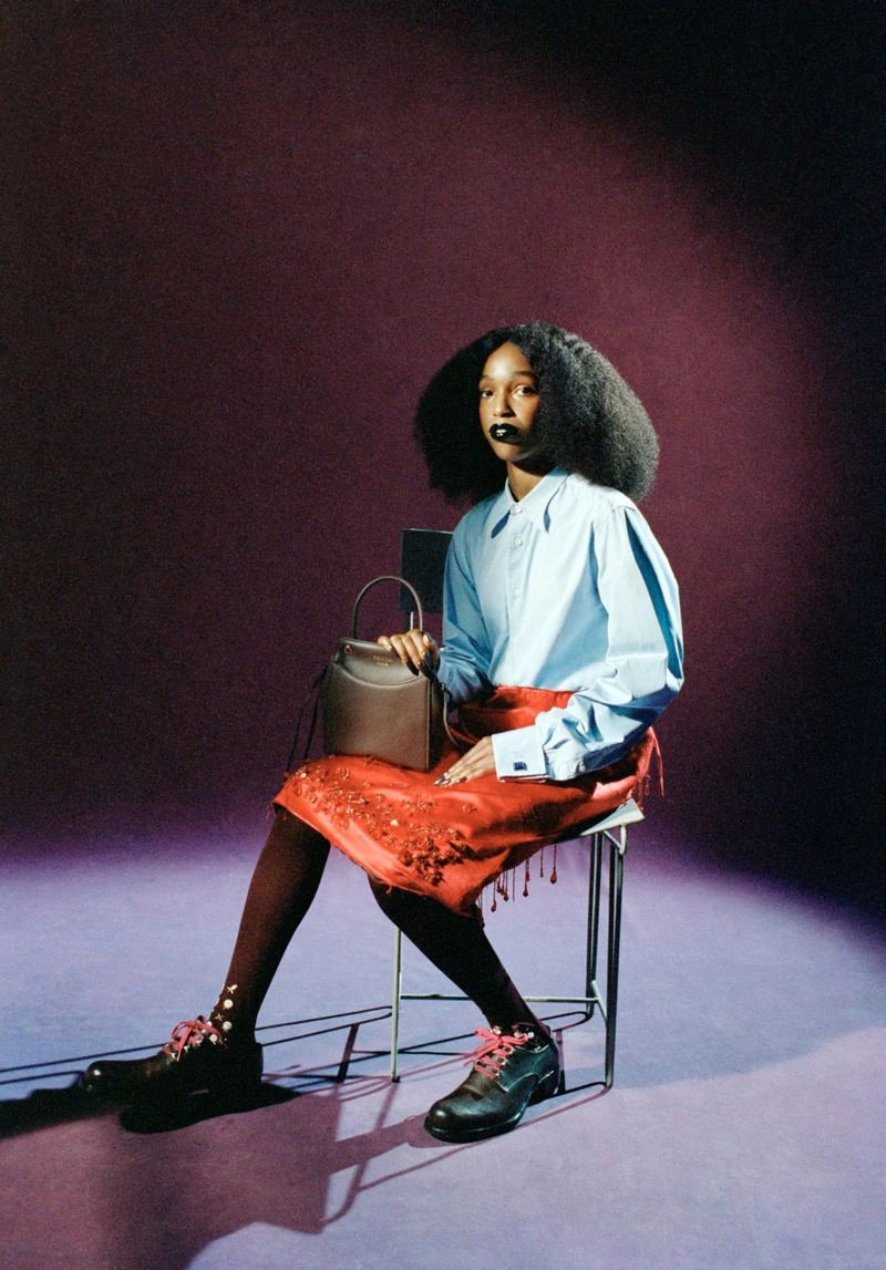 John Glacier seated on a metal chair wearing a light blue shirt and red skirt holding a handbag for i-D Magazine
