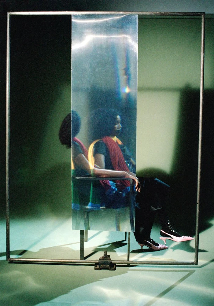 Portrait of John Glacier seated behind a vertical reflective strip within a metal frame for i-D Magazine by Camille Vivier.