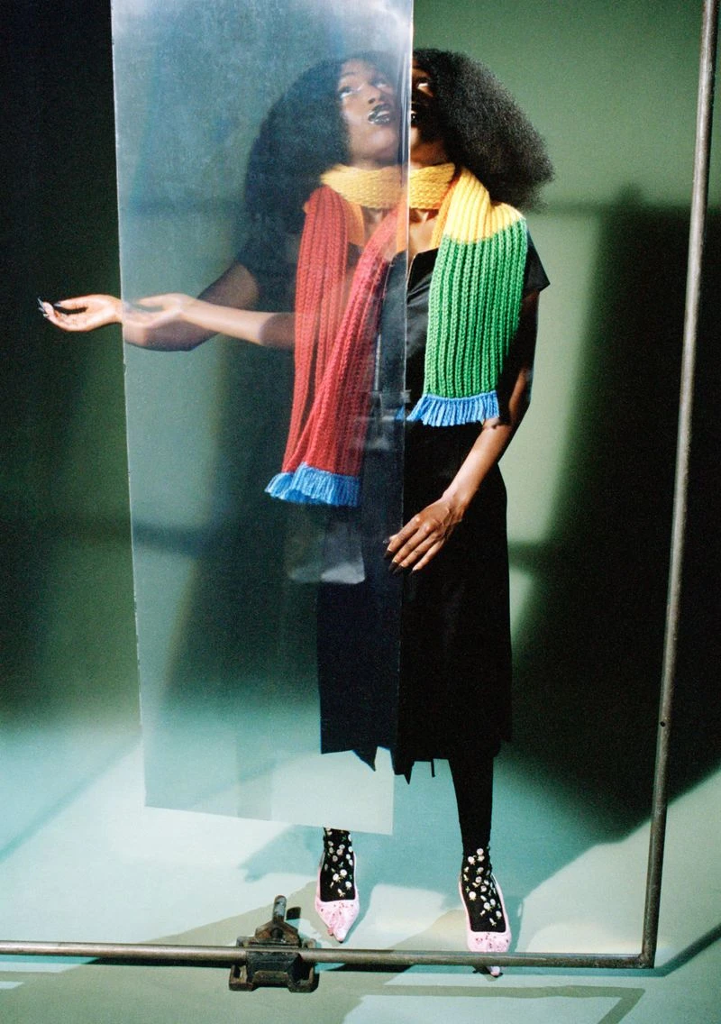 Portrait of John Glacier wearing a striped knit scarf and dark dress behind a glass panel for i-D Magazine
