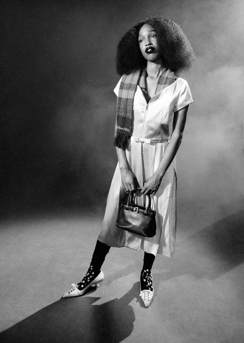 Black and white portrait of John Glacier wearing a belted dress and scarf, holding a handbag in a studio