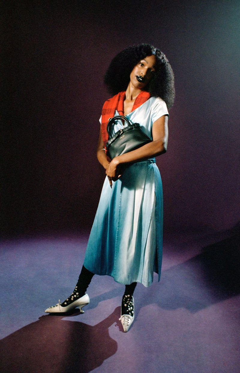 John Glacier in a light blue dress with a red scarf and black handbag holding a dark pose against a dark purple studio backdrop.